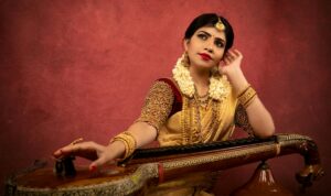 Elegant Indian woman in traditional wear posing with a Veena against a red backdrop.