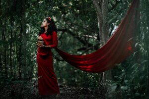 A woman in a long red dress poses elegantly in a lush forest setting, holding a woven basket.
