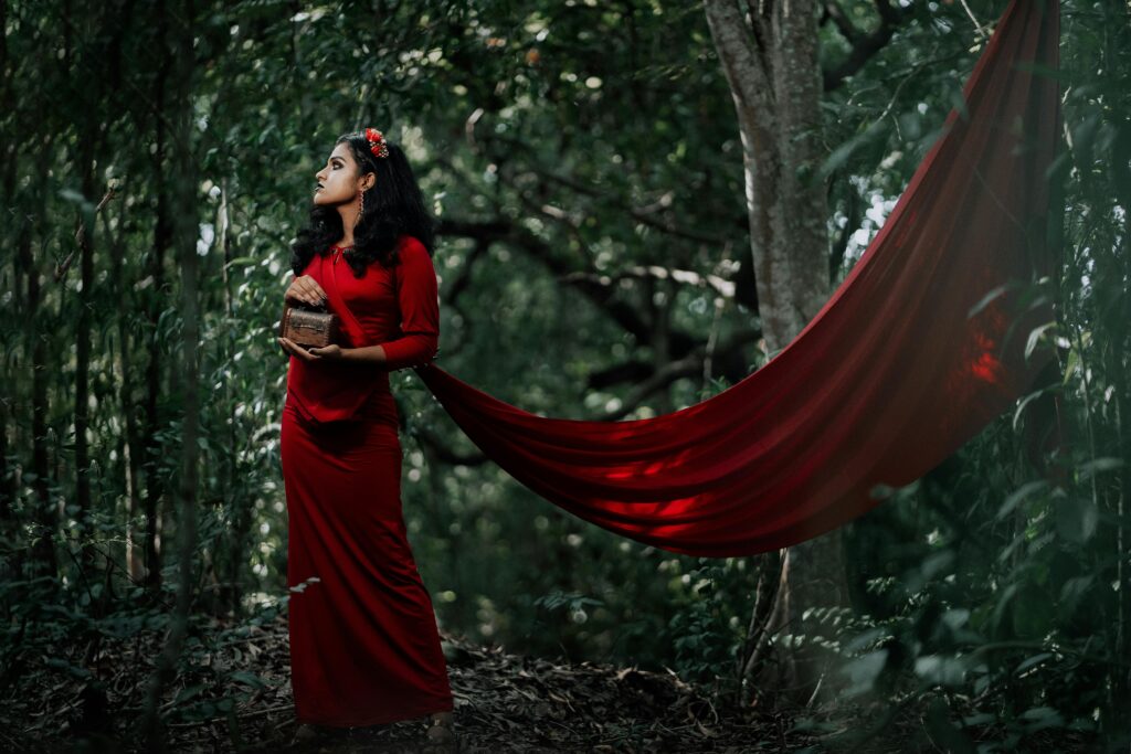 A woman in a long red dress poses elegantly in a lush forest setting, holding a woven basket.